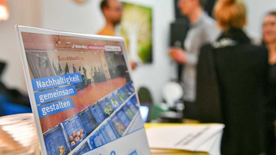 Ein Aufsteller "Nachhaltigkeit gemeinsam gestalten", dahinter Menschen, die sich unterhalten