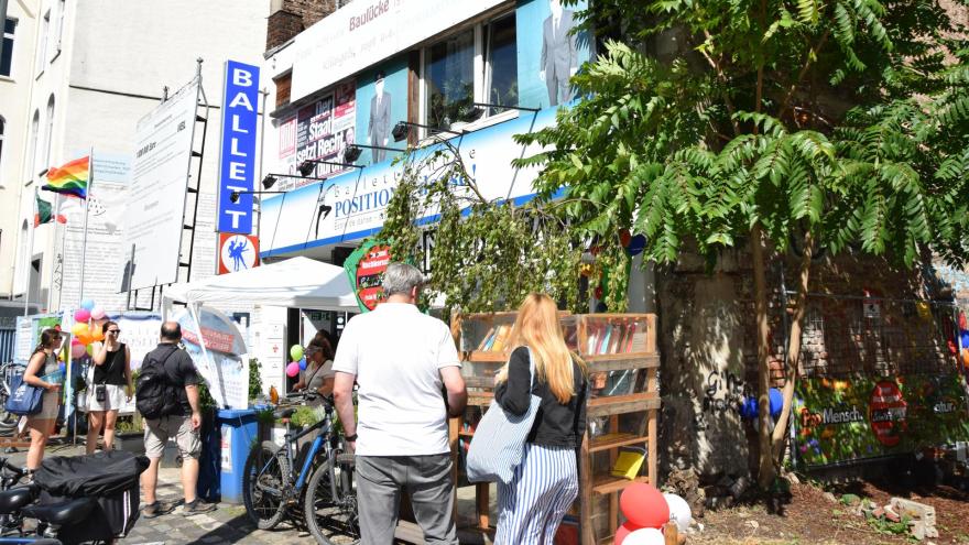 Auf dem Bild sind mehrere Personen zu sehen, die vor dem Haus der Initiative "Schmitzundkunzt" stehen. Sie schauen sich einen Bücherschrank und einen Stand zum Jeans-Recycling an.
