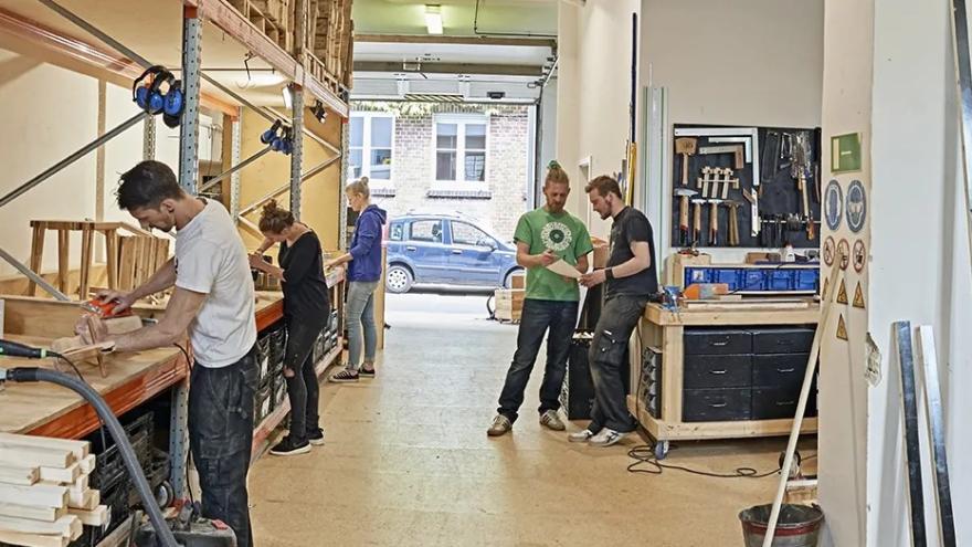 Im Bild sind mehrere Menschen zu sehen, die in einer Werkstatt am werkeln sind.