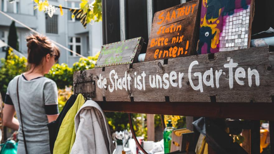 Das Bild zeigt eine Gartenaufnahme, eine Person ist mit dem Rücken zur Kamera zu sehen. Auf einem Holzbalken steht der Slogan "Die Stadt ist unser Garten" in selbstgemalter weißer Schrift.