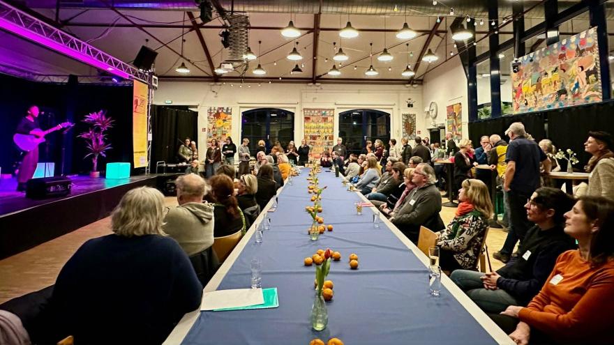 An einer langen Tafel sitzen viele Personen, die auf eine Bühne mit Live-Musik blicken. 