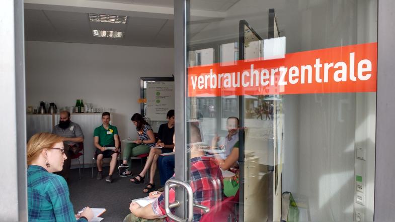 Geöffnete Tür beschriftet mit Verbraucherzentrale bietet Blick in den Raum, in dem Leute im Kreis sitzen