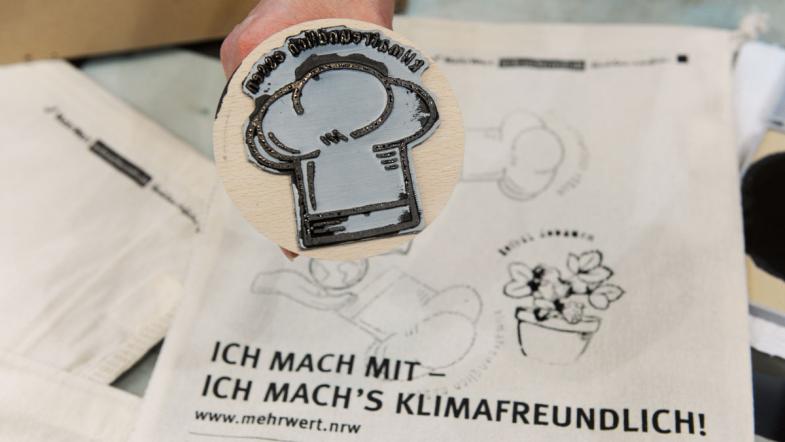 Eine Hand hält einen Stempel, darunter liegt ein Baumwollbeutel