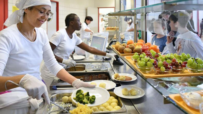 Zwei Mitarbeiterinnen des Mensapersonals verteilen Essen auf Teller und händigen diese an zwei Schüler aus.