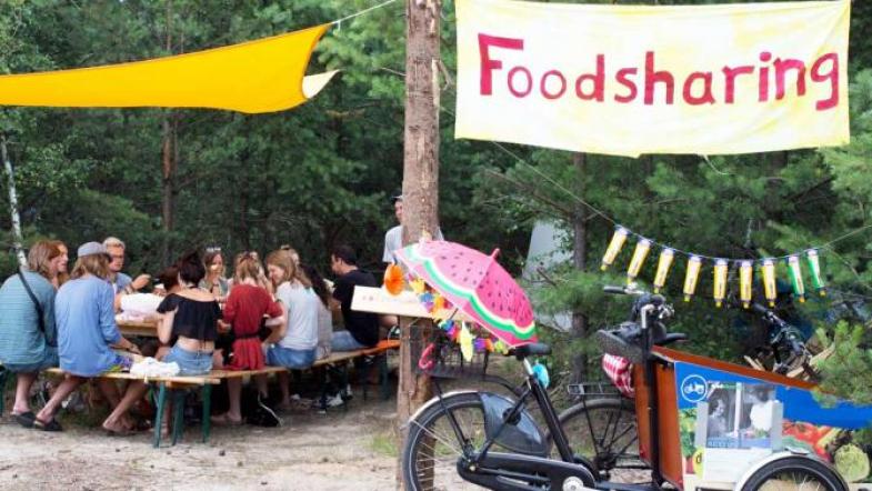 Menschen sitzen gemeinsam im Freien an einem Tisch, neben ihnen hängt ein "Foodsharing"-Banner