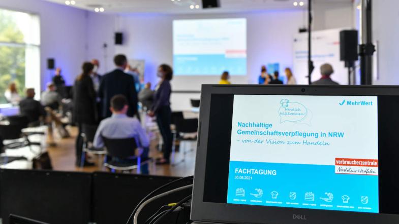 Ein Laptop mit Stream der Veranstaltung, im Hintergrund sieht man Teilnehmer der Fachtagung