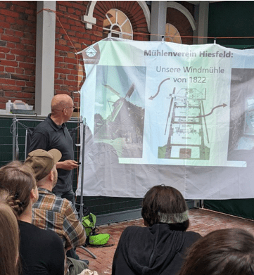 Auf dem Bild sind mehrere Personen auf Bänken mit dem Rücken zum Bildbetrachter zu sehen. Sie schauen auf eine Leinwand. Das Bild enstand im Rahmen des Stammtischtreffens der Initiative.