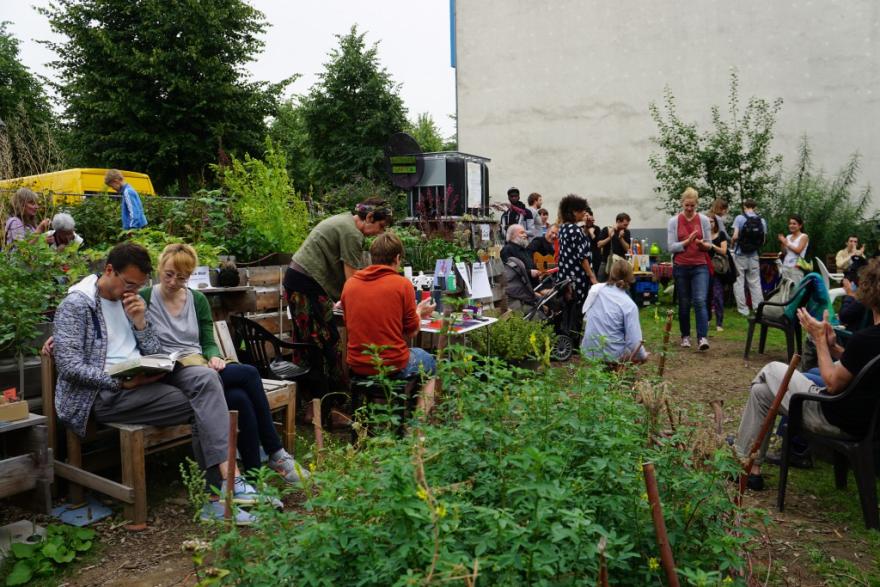 Auf dem Bild sind ungefähr 20 Personen im Gemeinschaftsgarten zu sehen. Sie sitzen im Grünen, unterhalten sich oder lesen. 